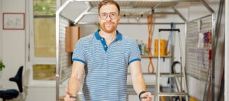 Oliver Schulz schiebt bei der Ergotherapie ein Trainingsgerät nach vorne. Er trägt ein blau-weiß-gestreiftes Poloshirt und schaut lächelnd in die Kamera. Im Hintergrund befindet sich ein nachgebauter Arbeitsplatz.