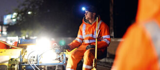 Ein Bauarbeiter arbeitet bei Dunkelheit an Bahnschienen. Er sitzt auf einem Arbeitsgerät und trägt orangene Warnkleidung sowie eine Kopflampe.